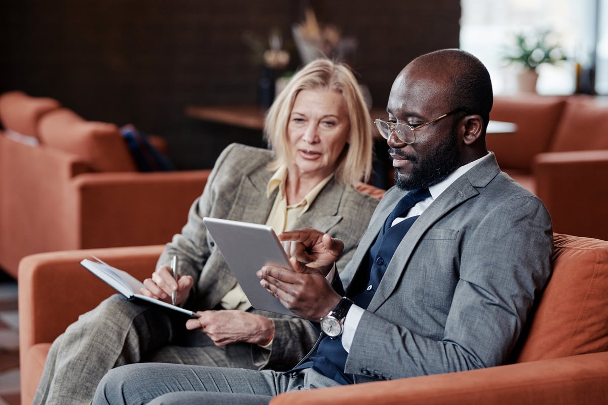A business owner pointing at a tablet screen while a colleague takes notes in a cafe meeting