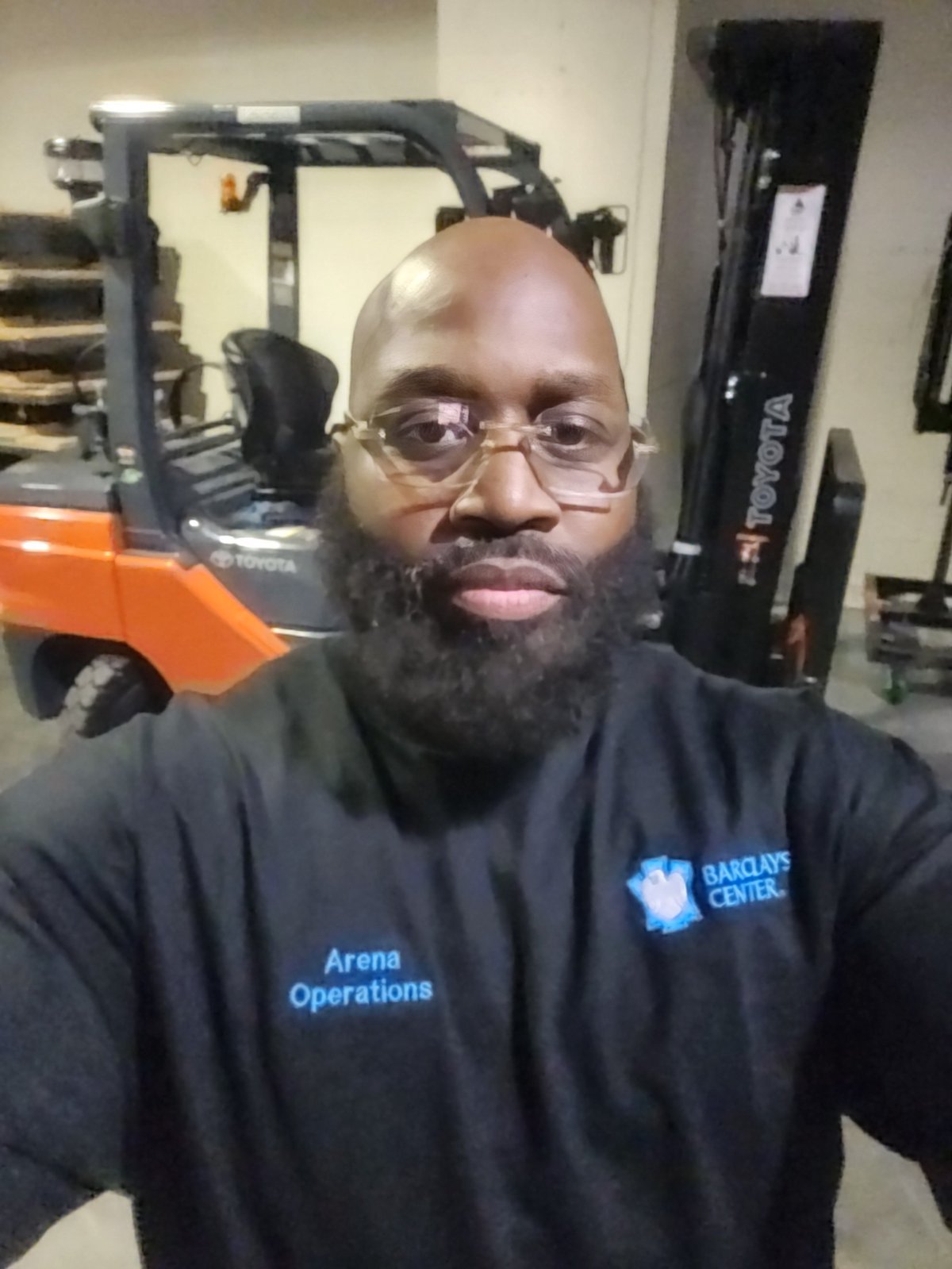 Justice Cephas wearing a Barclays Center Arena Operations shirt and glasses in an event warehouse with a forklift in the background