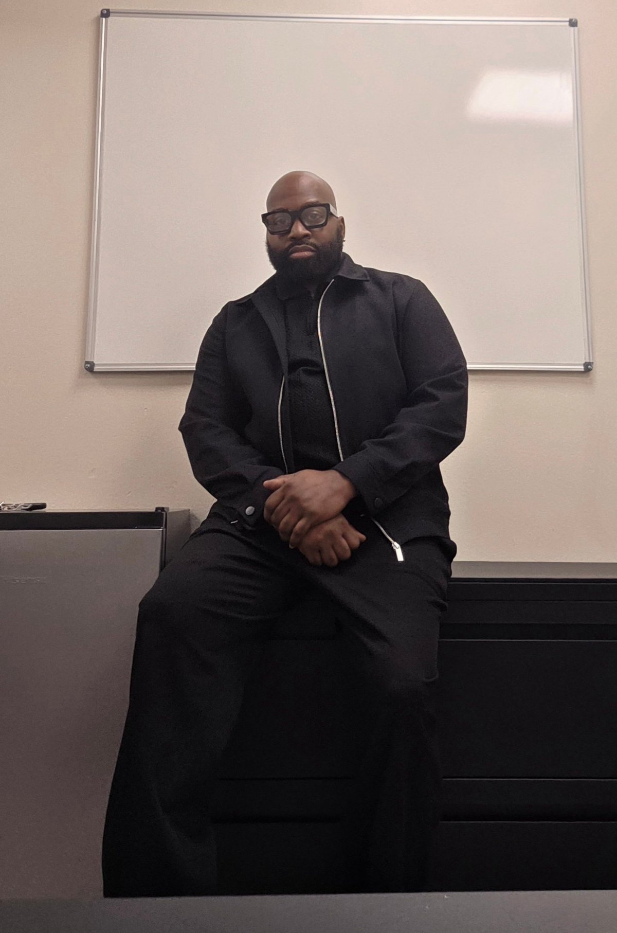 Justice Cephas, owner and operator of Kapital Enterprises, seated in front of a whiteboard in a black jacket and glasses